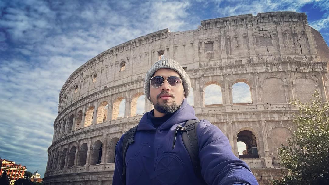 Elio in sunglasses and a beanie in front of the Colosseum.